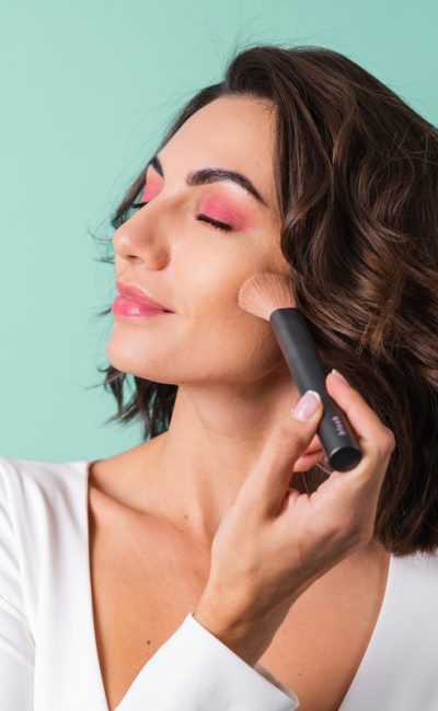 Young woman in a white evening dress on a light green background with bright pink makeup holding a blush brush